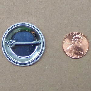 May include: The back of a round button with a silver metal backing and a pin on the back. The button is about the size of a US penny.