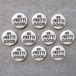 May include: A collection of ten circular white pin-back buttons arranged in three rows. Each button features the black text BE PRETTI GOOD printed in a bold, sans-serif font. The buttons have a glossy finish and a smooth, rounded edge design.