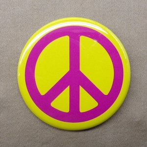 May include: A circular pin-back button featuring a bright yellow background with a bold, magenta-colored peace symbol centered on the surface. The design is clean and graphic, with a smooth, glossy finish covering the entire round face of the item.