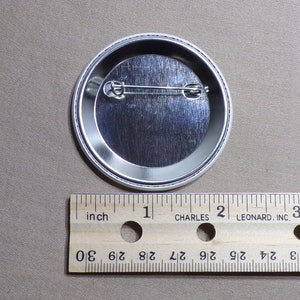 May include: The back of a silver button with a pin on the back. The button is 1 inch in diameter.