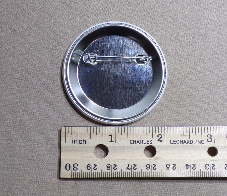 May include: The back of a silver button with a pin on the back. The button is 1 inch in diameter.