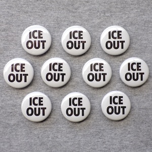 May include: A collection of ten white round buttons with the words "ICE OUT" printed in bold black letters. The buttons are arranged on a gray surface.