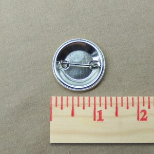 May include: The back of a silver button with a pin attached. The button is 1 inch in diameter.