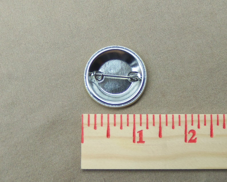 May include: The back of a silver button with a pin attached. The button is 1 inch in diameter.