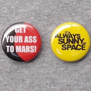 May include: Two round pinback buttons. One button is red and black with the text "GET YOUR ASS TO MARS!". The other button is yellow with the text "IT'S ALWAYS SUNNY IN SPACE".