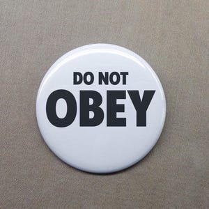 May include: A circular pin-back button with a white background and bold black text that reads DO NOT OBEY. The design features the word DO NOT in a smaller font size above the larger, prominent word OBEY, centered on the smooth surface.