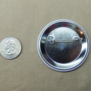 May include: The back of a silver button with a pin and a metal clasp. The button is about the size of a US quarter.