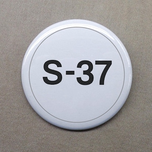 May include: White round button with black text that reads "S-37".