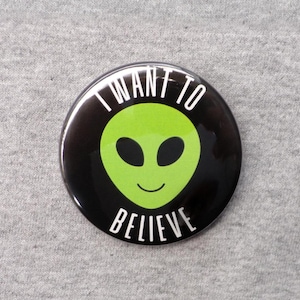 May include: A round black button with the text "I WANT TO BELIEVE" in white. The button features a green alien face with large black eyes and a smiling mouth. The button is a novelty item.