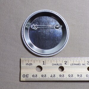 May include: The back of a silver button with a safety pin attached. The button is 1 inch in diameter.