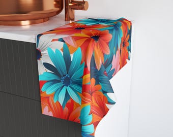 Puede incluir: Una toalla de mano con un estampado floral colorido. El diseño presenta grandes flores en tonos de azul, naranja, rojo y blanco. La toalla está colocada sobre una superficie blanca, mostrando el patrón audaz.