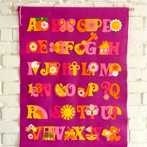 May include: A purple felt alphabet banner with colorful felt letters and illustrations. The letters are in a retro style with a variety of designs, including a house, a flower, a guitar, and a sun.