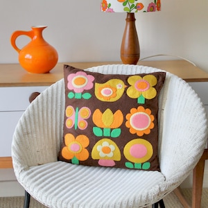 May include: A brown decorative pillow with colorful felt flower and butterfly appliqués sits on a white wicker chair. An orange pitcher and a wooden lamp with a floral lampshade are in the background. The pillow has a retro, vintage aesthetic.