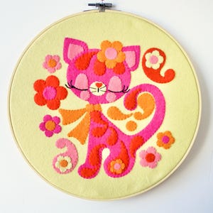 May include: A pink felt cat with orange and yellow accents, embroidered with a flower crown, on a yellow felt background. The cat is surrounded by flowers and swirls.