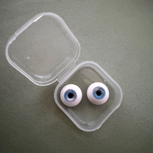 May include: Two spherical artificial eyes with light blue irises and black pupils, resting inside a small, square, clear plastic storage container with a hinged lid that is open and positioned to the upper left of the base.