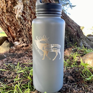 Peut inclure: Bouteille d'eau en acier inoxydable grise avec un couvercle noir et une poignée de transport. La bouteille est gravée au laser d'un motif d'élan avec des montagnes en arrière-plan et le texte "Be the Wild".