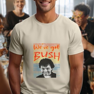 May include: Cream-coloured t-shirt featuring the text "We've got BUSH" in orange and red, with an image of a smiling man. The shirt is worn by a person in a social setting.