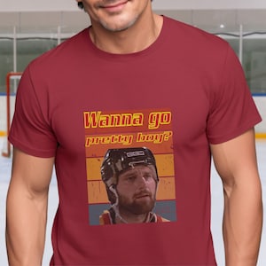 May include: Red t-shirt featuring a retro-style graphic of a hockey player wearing a helmet and a beard. The text "Wanna go pretty boy?" is above the image.
