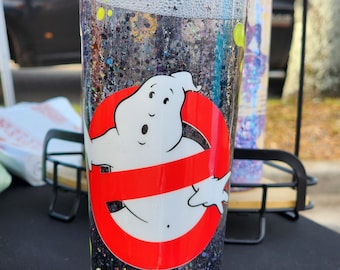 May include: A clear tumbler with a black glitter base and a Ghostbusters logo. The logo features a white ghost inside a red circle with a red slash. The tumbler has a clear lid and is filled with glitter and bubbles.