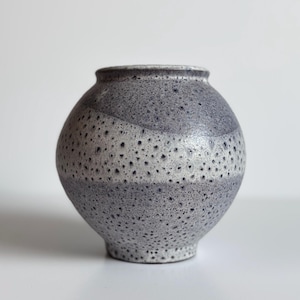 Peut inclure: Un vase en céramique sphérique avec une finition tachetée grise et blanche. La surface présente des bandes horizontales de différentes nuances de gris, avec un motif dense de taches sombres sur tout le corps. Le vase a un col étroit et une petite base.