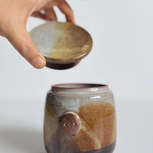 May include: A ceramic jar with a removable lid, featuring a mottled brown, tan, and grey glaze. A small circular medallion is attached to the side of the jar, embossed with the name JACK in capital letters. The jar has a smooth, rounded shape.