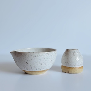 May include: A ceramic mixing bowl with a pouring spout and a small matching ceramic vessel. Both items feature a white speckled glaze on the upper portion and a natural, unglazed tan clay base, showcasing a simple, functional design for kitchen use.