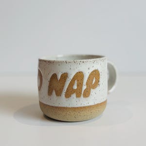 Peut inclure: Une tasse en céramique avec un émail blanc moucheté sur la partie supérieure et une base beige unie. Le mot NAP est gravé en grandes lettres texturées de couleur beige sur le devant. La tasse présente une anse arrondie et une finition mouchetée.