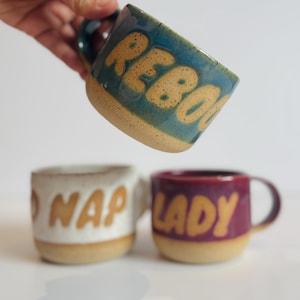 May include: Three ceramic mugs with speckled tan bases and coloured upper sections. The top mug is blue with the text REBOOT. The bottom left mug is white with the text NAP LADY. The bottom right mug is maroon with the text LADY.