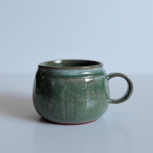 May include: A rounded ceramic mug featuring a mottled sage and forest green glaze with a smooth, glossy finish. The mug has a wide, slightly tapered body, a thick circular handle on the right side, and a visible reddish-brown clay base.