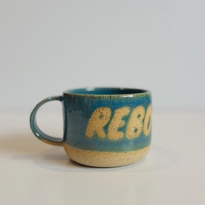 Peut inclure: Une tasse en céramique avec une finition mouchetée, présentant un design bicolore avec une partie supérieure bleue et une partie inférieure beige. Le mot REBO est embossé en grandes lettres beiges arrondies sur la surface bleue de la tasse.