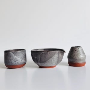 May include: A set of three ceramic vessels featuring a speckled grey and black glaze on the upper portions and a solid terracotta-coloured base. The collection includes a small cup, a wider bowl with a pouring spout, and a tapered vase-like container.