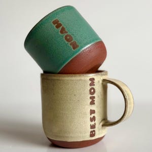 May include: Two handmade ceramic mugs. One is teal with the name "NOAH" in brown lettering. The other is beige with "BEST MOM" in brown lettering. Both mugs have a brown base and are stacked on top of each other.