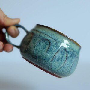 May include: A ceramic mug featuring a glossy, mottled teal and blue glaze with a series of vertical, arched indentations around the exterior. The mug has a curved handle and a dark, reddish-brown base, showcasing a handcrafted, textured finish.