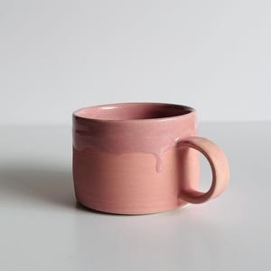 May include: A ceramic mug featuring a two-tone design with a glossy, light pink glaze on the upper portion that drips down over a matte, coral-pink base. The mug has a wide, cylindrical body and a curved handle on the right side.