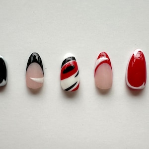 May include: Five oval-shaped false nails with different designs. The nails are white with black and red accents. One nail has a black and white swirl design, one has a red and white swirl design, one has a red and white stripe design, one has a black tip with a white line, and one has a black tip with a white line.