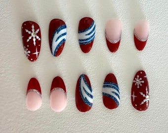 Puede incluir: Un conjunto de diez uñas artificiales con diversos diseños. Las uñas presentan una base rojo oscuro con diseños de copos de nieve y rayas blancas, y algunas tienen un acento plateado brillante. Otras uñas son de color rosa claro con una punta roja y plateada.