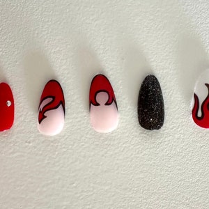 May include: Five different fake nails with various designs. One nail is red with three silver rhinestones, one is white with a red and black flame design, one is white with a red and black abstract design, one is black with glitter, and one is white with a red flame design.