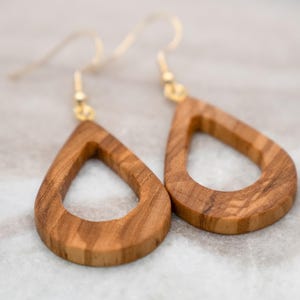 May include: A pair of teardrop-shaped earrings. The earrings are made of light brown wood with visible grain patterns. They have gold-colored ear wires. The earrings are displayed on a light-colored, blurred background.