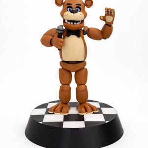 May include: A brown bear figurine with a black top hat, bow tie, and microphone. The bear is standing on a black and white checkered platform. The bear has a tan chest and blue eyes, with one hand raised in a wave.