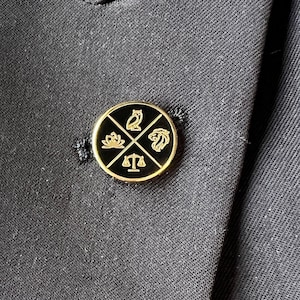 Puede incluir: Un pin de solapa negro y dorado con un diseño circular. El círculo presenta una balanza de la justicia, un pájaro, una flor y un pez. El texto "STA" está escrito debajo de la balanza de la justicia.