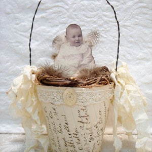 Könnte beinhalten: Ein dekorativer Korb im Vintage-Stil mit einem Babybild, Federn und einem Drahtgriff. Der Korb besteht aus einem papierbezogenen Topf mit handschriftlichem Schriftzug und Spitzentrimm. Die Farbpalette ist cremefarben und braun.
