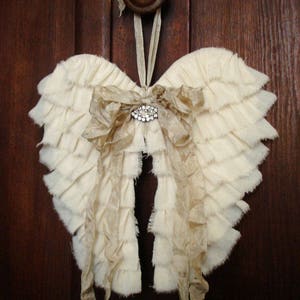 May include: A pair of white fabric angel wings with a brown ribbon bow and a rhinestone embellishment. The wings are hanging from a brown door knob.