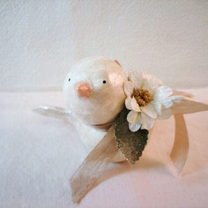 May include: A small, round, off-white bird figurine with a smooth, glossy finish. The bird has two tiny black dot eyes and a pale pink beak. A white fabric flower with a yellow center, a green leaf, and a beige ribbon are attached to its side.