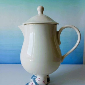 Peut inclure: Une théière en céramique blanche avec un couvercle et une anse. La théière a deux pieds en forme de chaussures bleues à pois blancs.