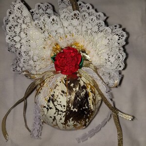 May include: A gold and white fabric ornament with a red rose and white lace trim. The ornament has a gold ribbon attached to the top.