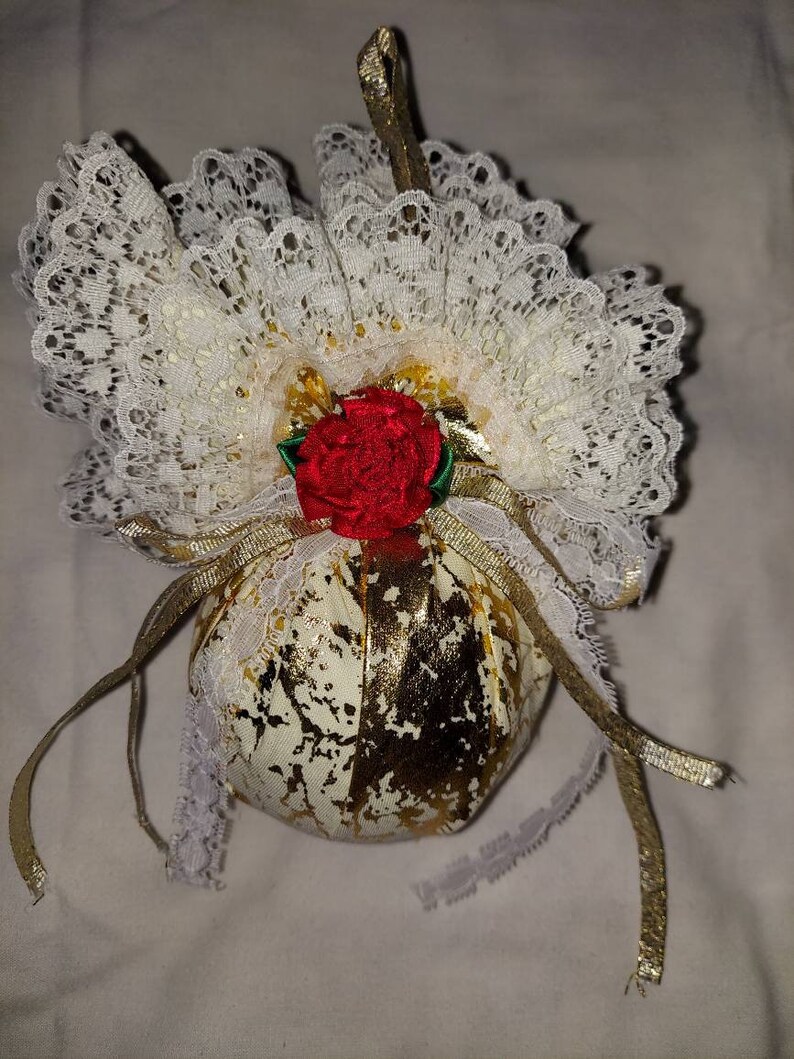May include: A gold and white fabric ornament with a red rose and white lace trim. The ornament has a gold ribbon attached to the top.