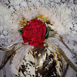May include: A white fabric ball with gold accents and a red rose on top. The ball is covered in white lace and has a gold ribbon tied around it.