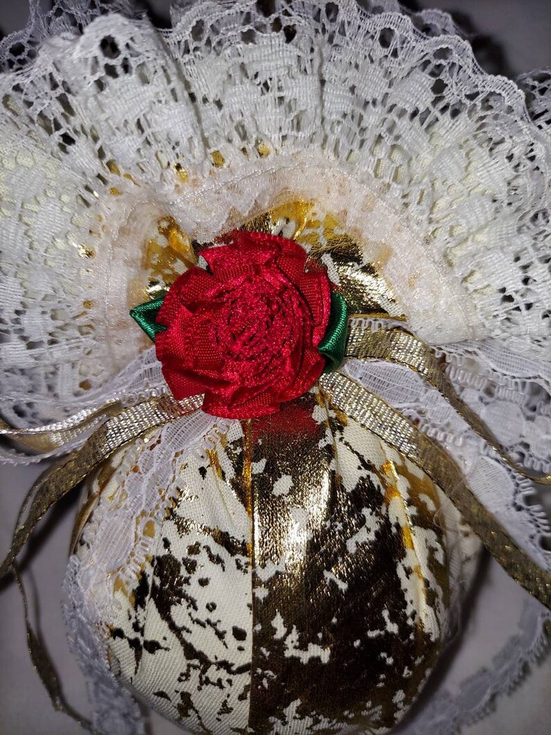 May include: A white fabric ball with gold accents and a red rose on top. The ball is covered in white lace and has a gold ribbon tied around it.