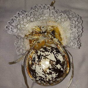 May include: A white and gold Christmas ornament with a lace trim. The ornament is shaped like a ball and has a gold ribbon attached to it.
