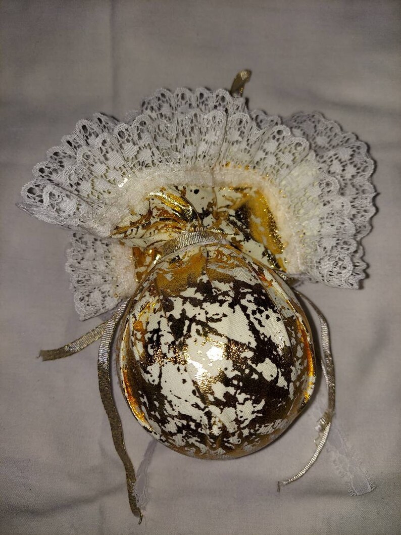 May include: A white and gold Christmas ornament with a lace trim. The ornament is shaped like a ball and has a gold ribbon attached to it.